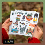 چند شیت استیکر با طرح‌های کمپینگ و طبیعت‌گردی شامل جنگل، قهوه کمپ، کفش کوهنوردی و المان‌های سفر در دستان یک زن در طبیعت پاییزی