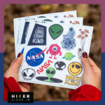 شیت استیکر چند طرحی شامل لوگوی ناسا، استیکر هکری Hack the Planet، شکلک لبخند (Smiley) با چشم‌بند، و شعارهای انگیزشی مانند It's cool to be kind در دست یک زن در طبیعت پاییزی
